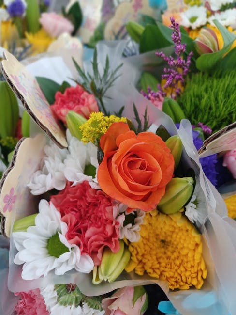A vibrant floral arrangement featuring a central orange rose surrounded by white daisies with green centers, pink carnations, yellow chrysanthemums, and sprigs of purple lavender, all wrapped in white